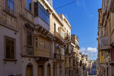 Sunlit row of ornate timber bay windows lines a descending street toward the harbor under a vivid blue sky.