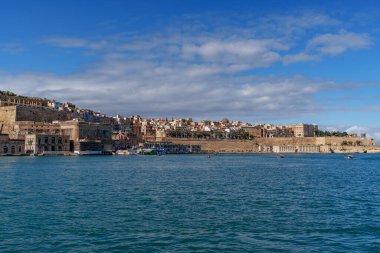 Limestone bastions and clustered buildings line the headland with a bright quay, seen over gentle harbor waters.