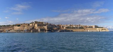 Wide coastal view showcases limestone bastions, quays and clustered buildings under bright sky with scattered clouds.