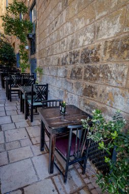 Row of small wooden tables and chairs lines a quiet stone lane with potted greenery and textured facade.
