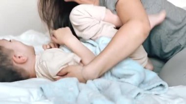 Anne, mavi bir battaniyeye sarılı küçük oğlunun ayaklarını gıdıklıyor. Anne ve çocuk evdeki rahat bir yatak odasında birlikte eğleniyorlar. Annenden mutlu çocuklar. Bebeklerin çocukluk fotoğrafları..