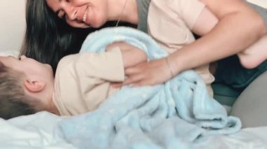 Anne, mavi bir battaniyeye sarılı küçük oğlunun ayaklarını gıdıklıyor. Anne ve çocuk evdeki rahat bir yatak odasında birlikte eğleniyorlar. Annenden mutlu çocuklar. Bebeklerin çocukluk fotoğrafları..