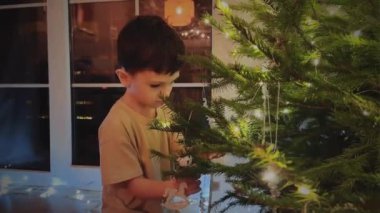 Küçük çocuk arka planda parlak kristallerle bir Noel ağacını süslüyor ve ağacın tadını çıkarıyor. Tatil ve Noel hazırlıkları, bir çocuğun yeni yıl ağacını süslemesi.