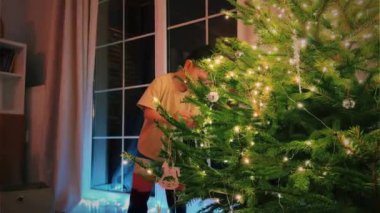 Küçük çocuk arka planda parlak kristallerle bir Noel ağacını süslüyor ve ağacın tadını çıkarıyor. Tatil ve Noel hazırlıkları, bir çocuğun yeni yıl ağacını süslemesi.