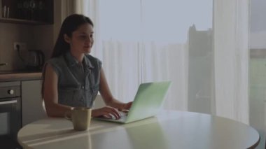 Neşeli, mutlu, pozitif ve güzel bir iş kadınının evde oturup bilgisayarını kullanması video konuşması ve mutfakta el sallaması..