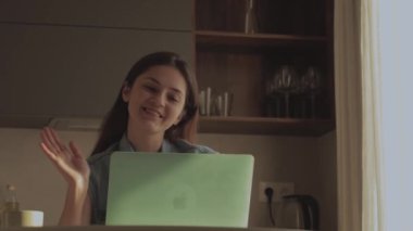 Neşeli, mutlu, pozitif ve güzel bir iş kadınının evde oturup bilgisayarını kullanması video konuşması ve mutfakta el sallaması..
