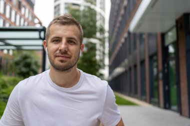 Sakallı, gülümseyen genç adam. Bir adam dizüstü bilgisayarda çalışıyor, video görüşmesinde konuşuyor, yan tarafa bakıyor, düz görünüyor. İnternette temiz havada uzaktan çalışıyor.