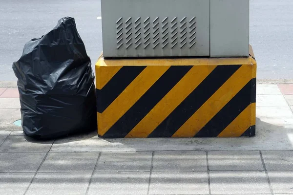 A yellow and black cement base warning sign of a telephone exchange booth with garbage bag on the sidewalk.