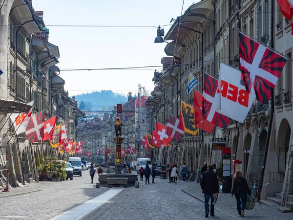 stock image View along Kramgasse, the main street in the historic city centre of Berne, Switzerland. Typical historic housing buildings, a historic fountain, and a richt decoration with flags.