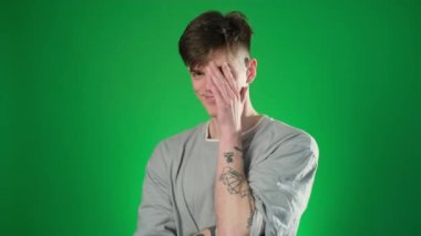 Gri tişörtlü adam utangaç ve gözlerini yeşil arka planda kapatıyor.
