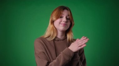 Kızıl saçlı beyaz kadın, hoşnutsuzluğu yansıtan bir şekilde giyinmiş, tarafsız yeşil zemine karşı mutsuz bir alkış tutuyor. İsteksiz alkışları memnuniyetsizlik hissi veriyor..