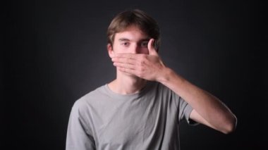 Melankolik beyaz bir adam, gündelik giyinmiş, ağzı eliyle kapatıyor, nötr siyah bir arkaplana karşı üzüntü veya içgözlem hissi veriyor. Bu jest bir sessizlik arzusu olduğunu gösteriyor..