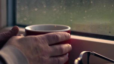 Sıcak içkisi olan kadın yağmurda pencereden dışarı bakar. 4K yakın çekim. Yavaş çekim seçici odaklanma.