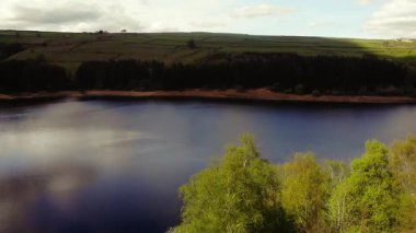Ağaçlar ve tarım arazileri, Peak District Ulusal Parkı 'ndaki gölü çevreleyen mavi gökyüzü geniş zoom insansız hava aracı seçici odak noktası.