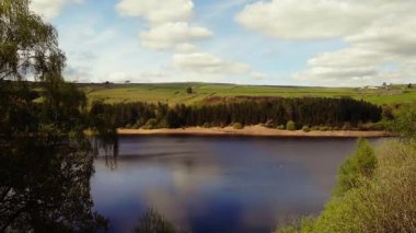 Ağaçlar ve tarım arazileri, Peak District Ulusal Parkı 'ndaki gölü çevreleyen mavi gökyüzü geniş zoom insansız hava aracı seçici odak noktası.