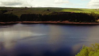 Ağaçlar ve tarım arazileri, Peak District Ulusal Parkı 'ndaki gölü çevreleyen mavi gökyüzü geniş zoom insansız hava aracı seçici odak noktası.