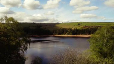 Ağaçlar ve tarım arazileri, Peak District Ulusal Parkı 'ndaki gölü çevreleyen mavi gökyüzü geniş zoom insansız hava aracı seçici odak noktası.