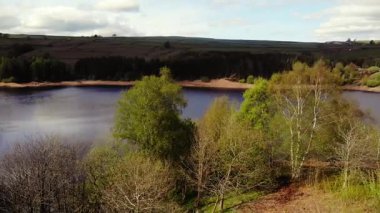 Ağaçlar ve tarım arazileri, Peak District Ulusal Parkı 'ndaki gölü çevreleyen mavi gökyüzü geniş zoom insansız hava aracı seçici odak noktası.
