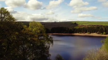 Ağaçlar ve tarım arazileri, Peak District Ulusal Parkı 'ndaki gölü çevreleyen mavi gökyüzü geniş zoom insansız hava aracı seçici odak noktası.