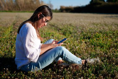 Telefonunu kontrol eden bir kadın, çiçek tarlasında oturuyor..