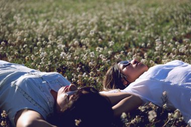 İki kadın, bir çiçek tarlasında elleri başlarının arkasında uzanmış, rahatlama işareti olarak..