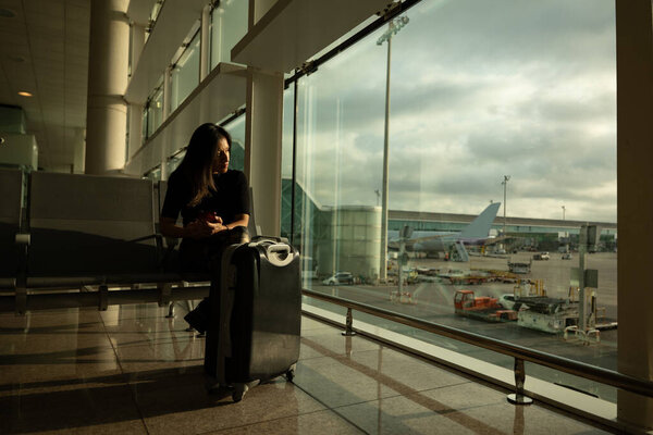 A woman in the airport boarding area, waits for her flight watching the landing strip on a cloudy day.