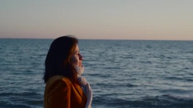 Gün batımında resmi giyinmiş ve fularlı bir kadın deniz kenarında yürüyor..