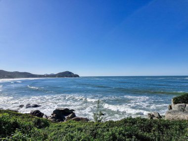 Dalgaların engebeli kayalara çarptığı, yemyeşil bitki örtüsüyle çevrelenmiş, mavi gökyüzünün altında, birkaç bulutla birlikte. Sahne bir sükunet, doğal güzellik, huzur, rahatlama ve doğaya kaçma hissi uyandırıyor..
