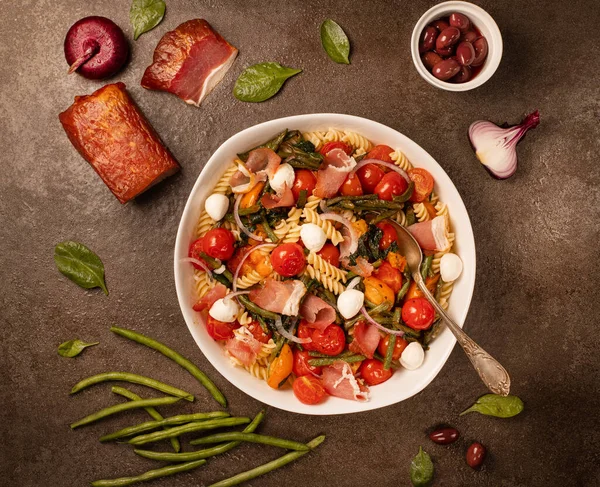 Salad with pasta, ham and vegetables in white bowl on gray stone backgroundtop view, copy space. Salad with pasta fusilli accompanied tomatoes, mozzarella, smoked ham and italian cheese