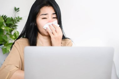 Hasta Asyalı kadın grip ve ateş yüzünden hapşırıyor ve öksürüyor. Masa başında kağıt mendil tutarken.