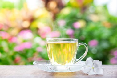 Sıcak çay bardağı ahşap masada, arka planda doğa bahçesi ve güneş ışığı var.