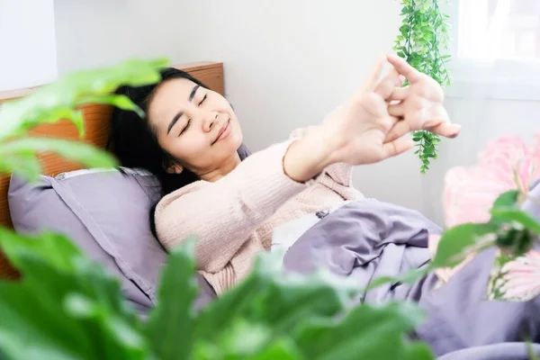 亚洲女人早上躺在床上醒来 张开双臂 笑容满面 喜见卧室里有新鲜的绿色植物 — 图库照片