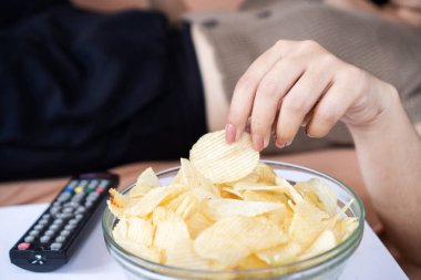 Kanepeye uzanmış patates cipsi yiyen, sağlıksız beslenen bir kadın. 