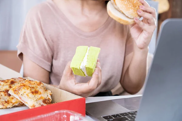 woman over eating fast food burger,  pizza and desserts at office desk, eating disorder concept