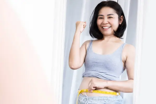 happy Asian woman success on dieting and weight loss measuring her waist with tape measure smiling in front of a mirror