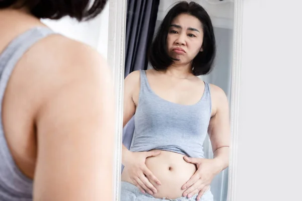 worried Asain woman checking her abdominal and looking at her fat belly in a mirror, weight gain concept