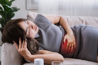 IBS, gıda zehirlenmesi ya da ishal yüzünden midesi ağrıyan Asyalı bir kadın. Elleri karnında ve masada bir şişe ilaç ile kanepede yatıyor.