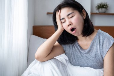 Asyalı kadın uykusuzluktan yorgun uyanıyor, esniyor ve yatakta bitkin düşüyor.