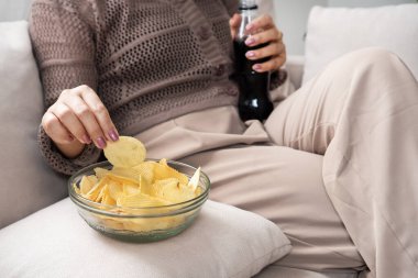 Patates cipsi yiyen, gazoz içen, duygusal beslenme davranışları gösteren ve yüksek miktarda şeker ve tuz içeren sağlıksız yemek seçenekleri sunan bir kadın.