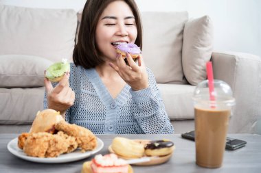 Asian woman showing signs of binge eating disorder while sitting at a table filled with fast food like burgers, fried chicken, donuts, cake, and sweet coffee drinks,  unhealthy eating habits 