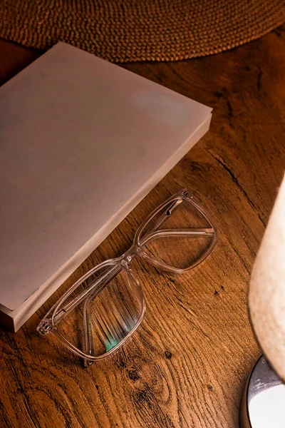 a pristine white-covered book, a stylish rustic hat, and classic glasses arranged on a warm oak table bathed in the soft glow of a table lamp.
