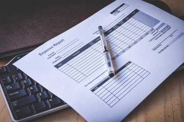 Siyah bir klavyenin ve kahverengi deri bir defterin üzerine yerleştirilmiş boş bir gider raporu formu, çam ağacından bir masanın üzerine..