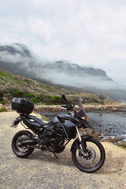 Kamp Koyu ve Hout Körfezi arasında manzaralı kıyı yolu boyunca siyah bir macera tarzı motosiklet park edilmiş. Arka planda 12 Havarinin ikonik dağ sıraları yükseliyor..