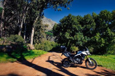 Beyaz macera motosikleti, sık yeşil ağaçlarla çevrili gölgeli toprak bir patikada park edilmiş ve Masa Dağı orman tepe örtüsünün içinden gözetliyor.