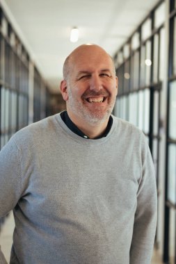 Kel, orta yaşlı, gri kazak giyen bir adam, cam panelli duvarları olan çağdaş bir ofis koridorunda dikilirken sıcak bir şekilde gülümsüyor.