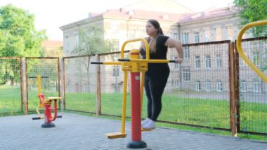 Siyah eşofmanlı şişman bir kız spor sahasında spor yapıyor. Genç bir kadın sıcak bir yaz gününde açık hava egzersiz makinesinde egzersiz yapıyor. Sağlıklı yaşam tarzı ve kilo kaybı.