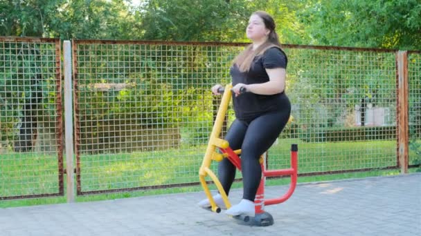 一个穿着黑色运动服的胖女孩正在运动场上健身 一个年轻的女人正在一个温暖的夏日里 在室外的运动机器上做运动 健康的生活方式和减肥 — 图库视频影像