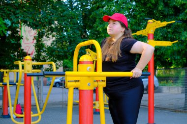 Siyah eşofmanlı şişman bir kız spor sahasında spor yapıyor. Genç bir kadın sıcak bir yaz gününde açık hava egzersiz makinesinde egzersiz yapıyor. Sağlıklı yaşam tarzı ve kilo kaybı.