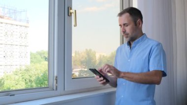 Profesyonel bir erkek, ofisteki bir akıllı telefondan mesaj yazıyor. Genç işadamı iş zamanı yönetim organizasyonu kavramı için kullanışlı mobil uygulamalar kullanıyor. Pencerenin yanında duruyor.