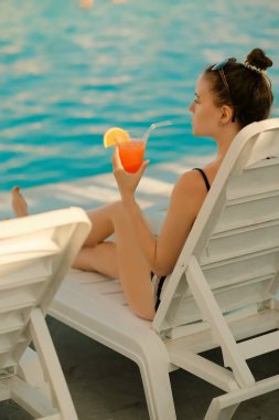 Girl in a sun lounger by the pool holding a cocktail in her hand clipart